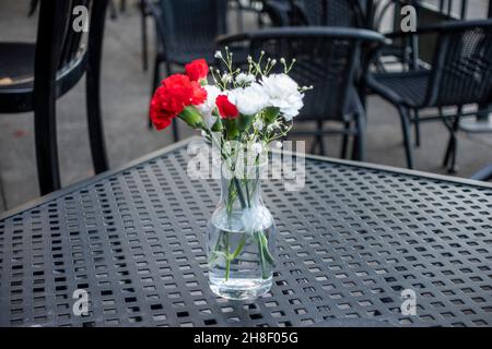 Side view of a glass vase filled with red and white carnations, on top of a black table in the outdoor seating area of a restaurant Stock Photo