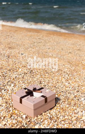 A box with a gift tied with a ribbon lies on the beach near the sea. Front view, vertical. Stock Photo