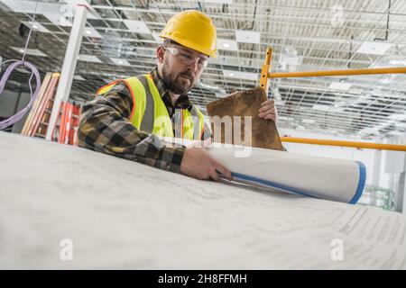 Construction Contractor Worker in His 30s Looking Inside Large Paper Building Blueprints. Construction Industry Theme. Stock Photo