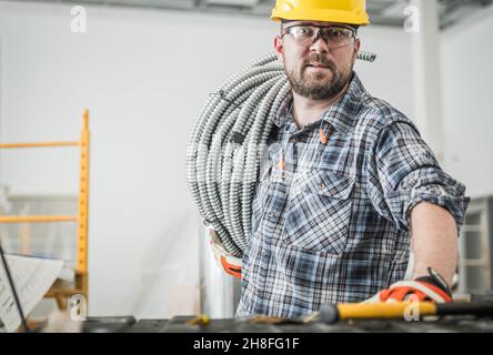 Caucasian Electrician Contractor Worker in His 30s with Metal Electric Cables Conduit Pipe on His Shoulder. Construction Site Theme. Stock Photo