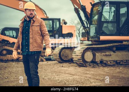 Ground Works Supervisor in His 30s Wearing Yellow Hard Hat and Staying Between Heavy Building Equipment Like Excavators. Stock Photo