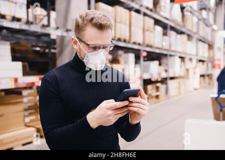 Young manager of store, man worker with protective mask using phone working in industrial factory or warehouse. Stock Photo