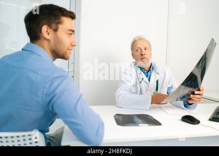 Mature male doctor radiologist explaining results of MRI scanning for handsome young man patient, showing snapshot with images, analyzing MRI scan. Stock Photo