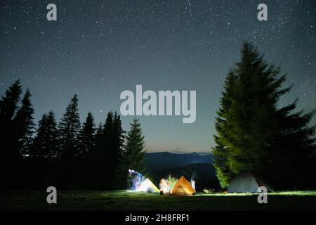 Bright illuminated tourist tents near glowing bonfire on camping site in dark mountain woods under night sky with sparkling stars. Active lifestyle an Stock Photo