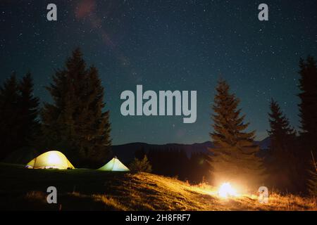 Bright illuminated tourist tents near glowing bonfire on camping site in dark mountain woods under night sky with sparkling stars. Active lifestyle an Stock Photo