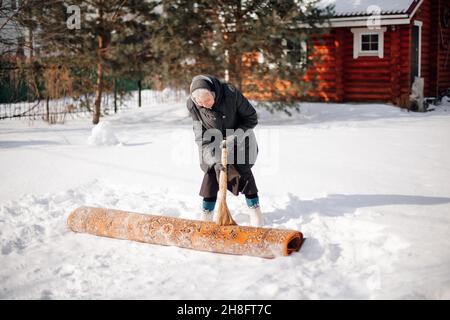 Caucasian woman in backyard. Elderly woman in scarf and felt boots cleans carpet in traditional way with help of fresh snow and broom, she is engaged Stock Photo