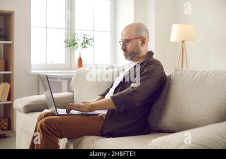 Serious adult man sitting on couch at home and working on modern laptop computer Stock Photo