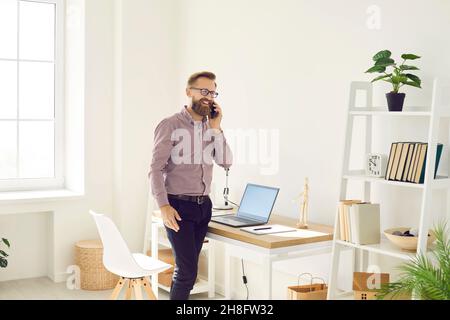 Happy businessman standing by office desk with laptop and talking on mobile phone Stock Photo