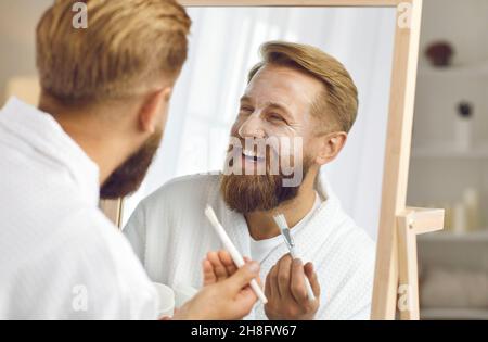 Close up mirror reflection of satisfied adult handsome bearded man who enjoys facial treatment. Stock Photo