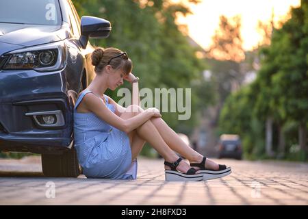 Young woman driver sitting beside her broken car waiting for help. Vehicle problems concept. Stock Photo