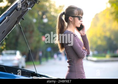 Young upset woman driver talking on mobile phone near a broken car with open hood waiting for help. Stock Photo