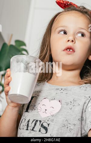 Little dark-haired girl 3 years old in red headband bakes apple pie in kitchen. Child holds measuring cup with flour. Children help on household Stock Photo