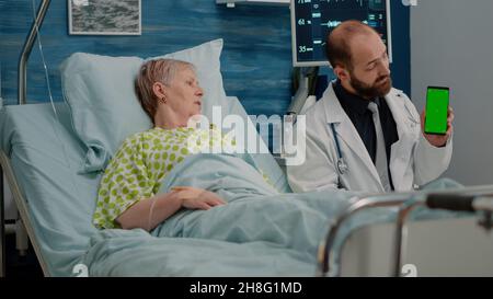 Doctor and sick patient looking at green screen on phone at nursing facility. Medical specialist vertically holding smartphone with chroma key for isolated template and mockup background Stock Photo