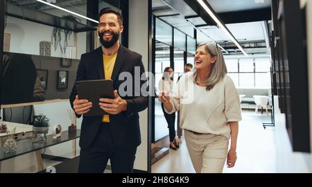 Laughing entrepreneurs walking together in an office. Two happy business colleagues sharing a laugh while having a discussion. Diverse businesspeople Stock Photo