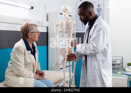 Specialist therapist doctor holding anatomical human skeleton arm explaining bones pain to senior retired woman patient during osteoporosis appointment in hospital office. Medicine concept Stock Photo