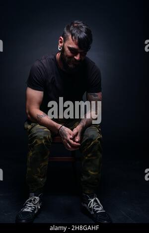 Brutal attractive bearded man with tattooed hands poses in black blank t-shirt and army pants seated on a chair Stock Photo