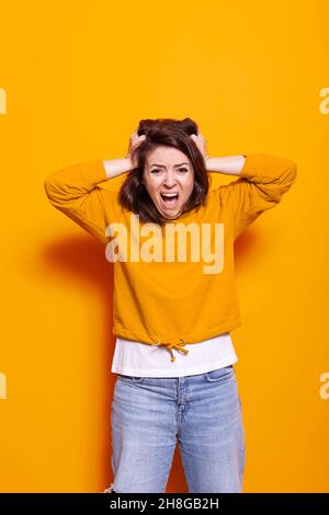 Desperate woman screaming and pulling hair with hands in studio. Young person with displeased and dramatic expression on face, feeling irritated while standing over orange background Stock Photo