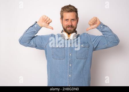Young smiling man in casual clothing wearing headphones pointing thumbs himself isolated over white background. Stock Photo