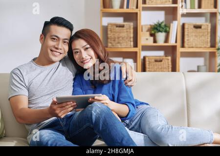 Portrait of positive young Asian couple sitting with tablet on sofa and embracing at home Stock Photo