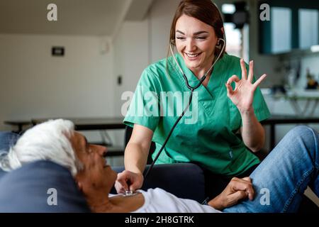 Health visitor using stethoscope to check health of a senior man, young nurse gesturing ok with hand during a home visit, home health care concept Stock Photo
