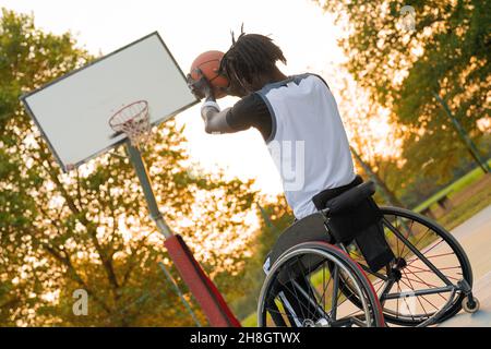 Young athlete on wheelchair throwing ball in basketball basket, sunset effect in background, social incluzion and sort diversity concept Stock Photo