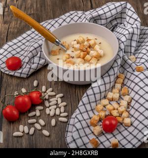 A bowl of semolina soup with toast cubes on top, on a wooden table with red cherry tomatoes and white dried beans Stock Photo