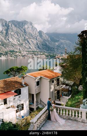 Groom hugs bride near the balustrade against the background of ancient houses on the coast of the bay Stock Photo