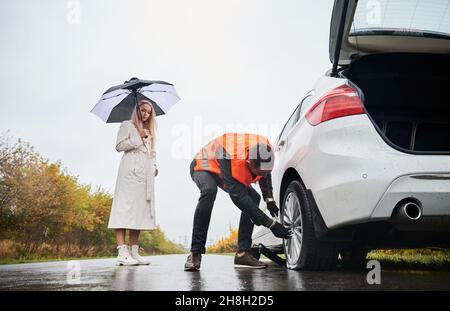 Male auto mechanic unscrewing lug nuts on car wheel while elegant woman holding umbrella. Young man in vest repairing woman automobile on the road. Concept of emergency road service. Stock Photo