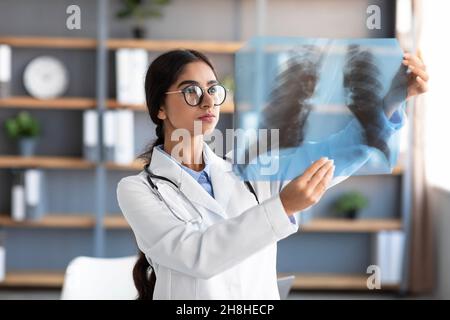 Busy serious millennial pretty indian woman doctor in white coat and glasses looking at lungs x-ray in clinic Stock Photo