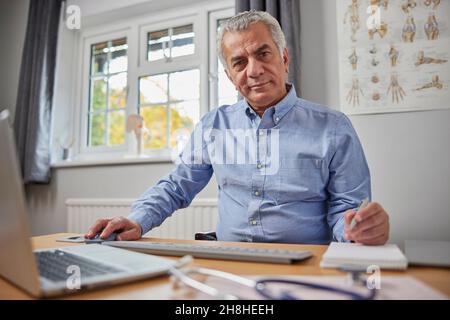Portrait Of Mature Male GP At Desk In Doctors Office Stock Photo