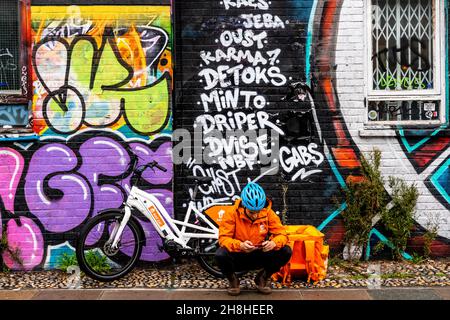 A ‘Just Eat’ Delivery Driver Sitting Down In The Street Using A Smart Phone, Shoreditch, London, UK. Stock Photo