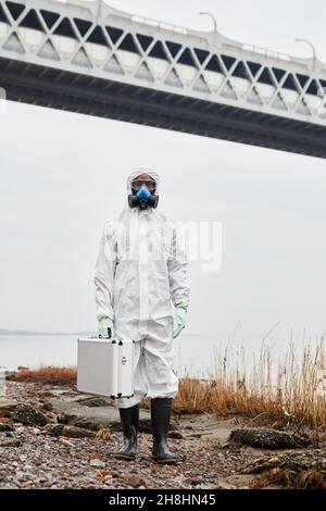 Vertical portrait of worker wearing hazmat suit walking by water outdoors and carrying samples case, industrial waste concept Stock Photo
