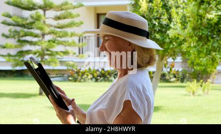 Senior female blonde businesswoman in hat uses tablet pc, digital tablet for business work or study in her own green garden. woman aged 50-55. The Stock Photo