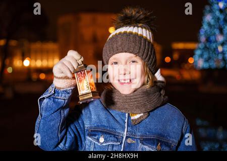 Small child waits for santa claus on street of small town during snowfall on christmas eve. Boy is holding lantern with burning candle. Children's hopes for magic and gifts. Winter holiday traditions Stock Photo