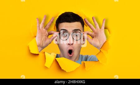 Shocked positive young european guy with open mouth, touches glasses, looks through hole in yellow paper Stock Photo