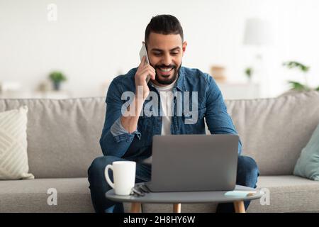 Work from home. Positive young Arab guy talking on mobile phone while using laptop computer indoors Stock Photo
