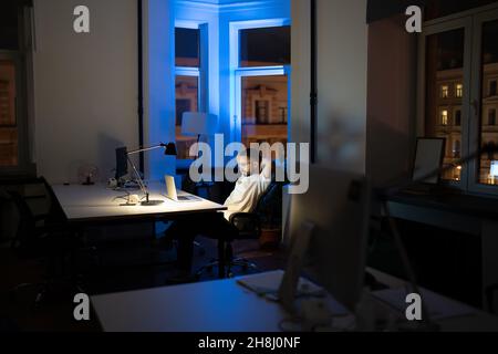 Happy calm man look at laptop hold hands behind head in night office satisfied with finished work Stock Photo