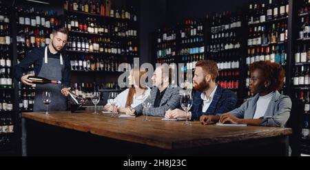 Group of European friends and African woman at wine tasting master class in restaurant, sommelier pours drink into decanter. Stock Photo