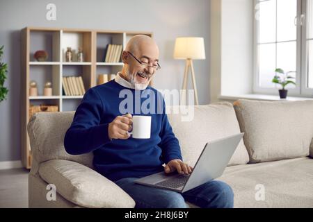 Happy senior man sitting on couch, using his laptop, drinking coffee and laughing Stock Photo