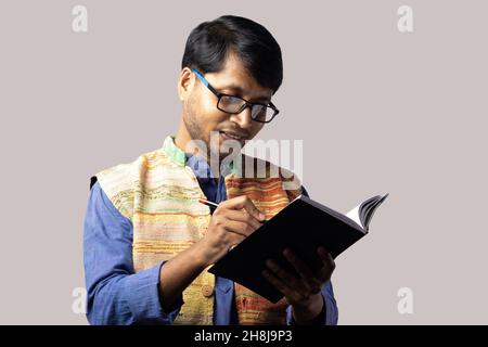 A young Indian male in ethnic dress writing with pen on notebook on gray background Stock Photo