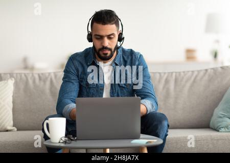 Arab man in headset using laptop pc, working online, having remote career, speaking to coworkers on web from home Stock Photo