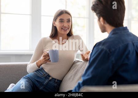 Happy emotional millennial caucasian woman with cup of drink sit on couch, gesturing, rest and communicating Stock Photo