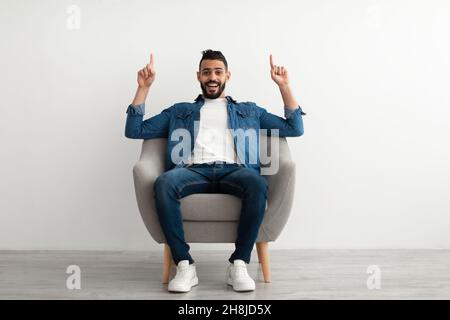 Full length of joyful young Arab guy pointing upwards with both hands, sitting in armchair against white studio wall Stock Photo