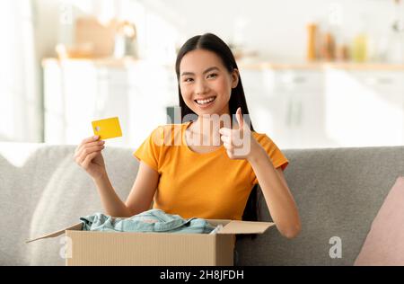 Home delivery. Young asian woman sitting on sofa with parcel, showing thumb up and credit card, smiling at camera Stock Photo