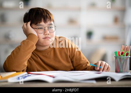 Asian schooler chubby boy dreaming about something while doing homework Stock Photo