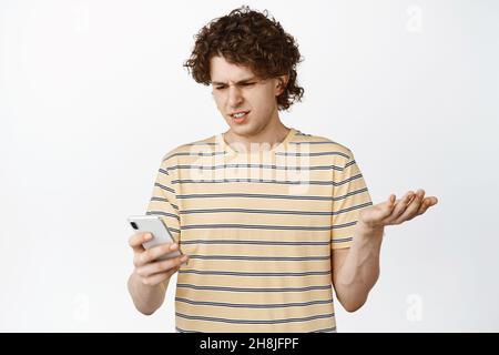 Image of curly guy staring confused at mobile phone, standing in t-shirt over white background Stock Photo