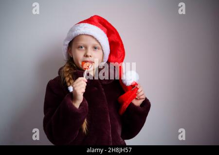 A Caucasoid Christmas girl in Santa's red cap and fur coat licks a lollipop against a light background. Stock Photo