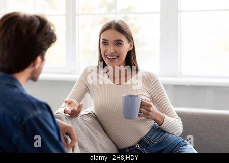 Cheerful young european husband and woman with cups of hot drink talk and spending weekend together Stock Photo