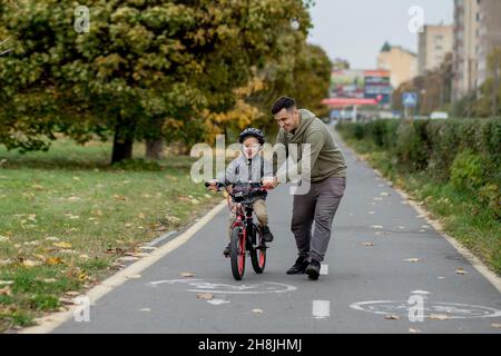 Father teaches his son to ride a bicycle on the bike path in the park. The father is holding a bicycle and the son is sitting on it. Stock Photo