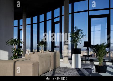 Front view of large panoramic window with incredible view on city and cozy grey sofa near beautiful green flowerpots. Concept of modern and stylish interior room. Stock Photo